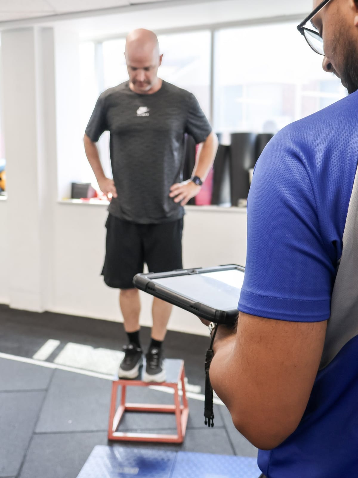 SportsFit physiotherapist recording force-plate results on a tablet while a patient steps down from a plyometric box onto dual force plates
