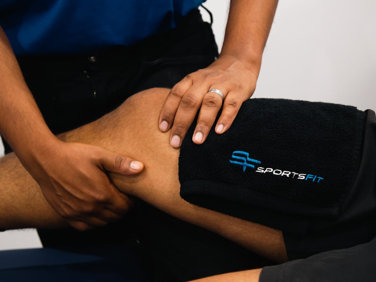 Close-up of a physiotherapist's hands assessing a patient's knee over a SportsFit-branded towel